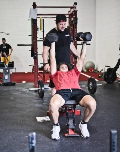 Iron Performance Gym Lifting Dumbbells with Help from Coach